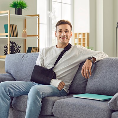 Un homme heureux portant une fronde orthopédique sur un bras blessé assis sur un confortable canapé à la maison, regardant la caméra et souriant. Blessure au bras, à la main, au poignet, à l'épaule ou au coude, concept de traitement conjoint en panne